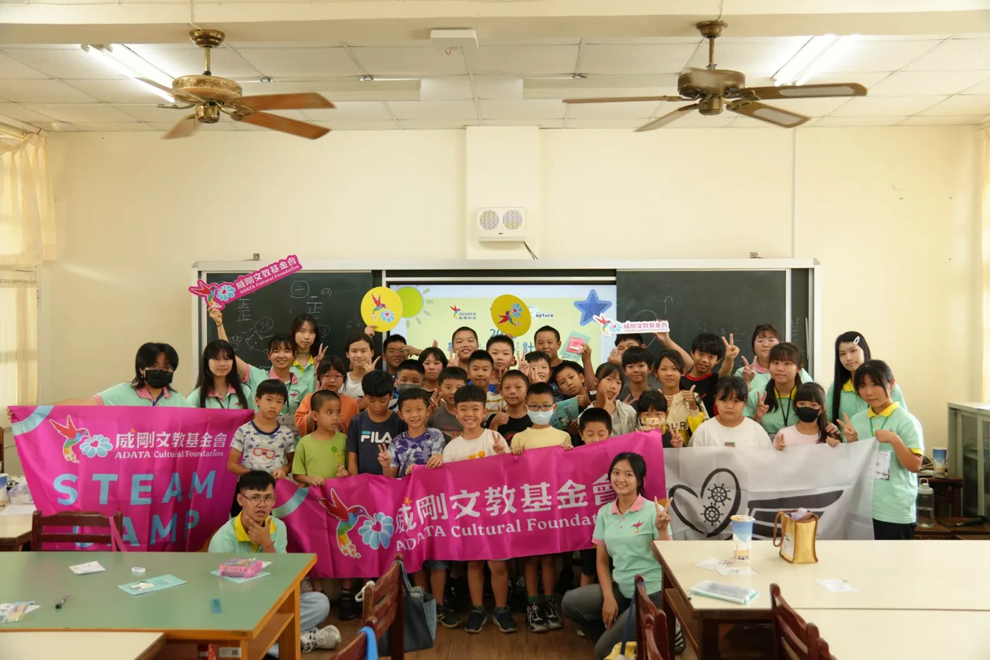 志工與孩子站在教室裡,舉著威剛文教基金會的 STEAM CAMP 旗幟與展心計畫的旗幟合照