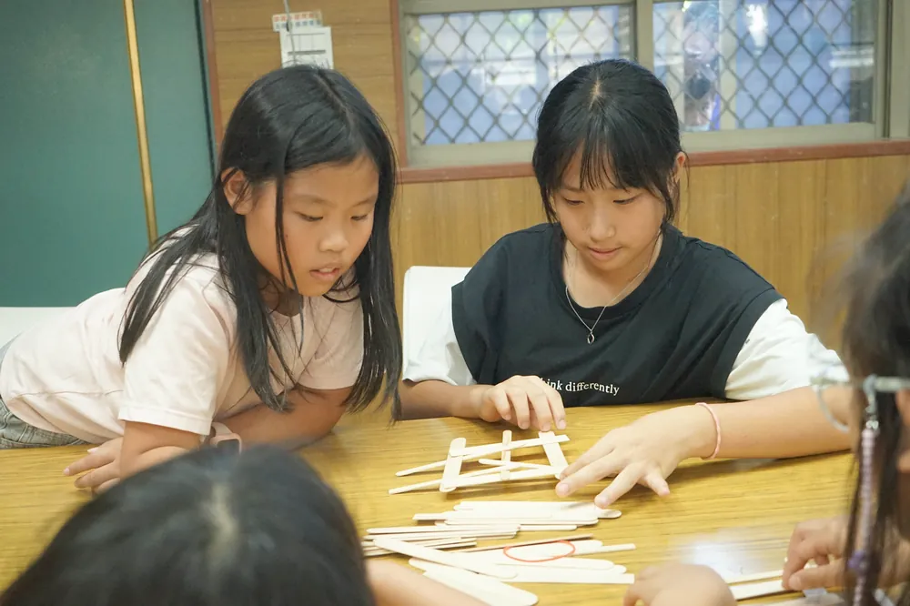 兩個孩子趴在桌邊，專心地用木棍搭橋
