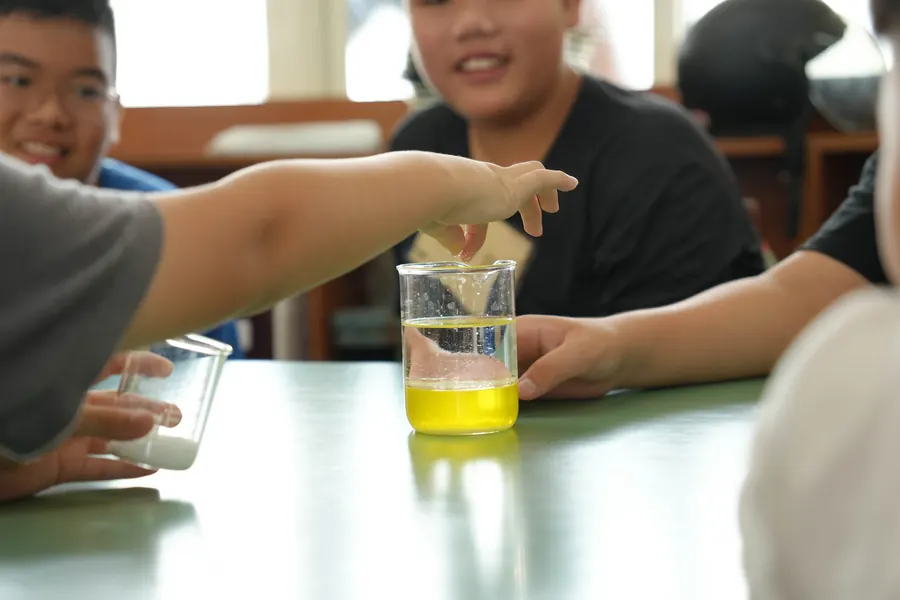 孩子把手伸向一杯黃色液體（加了色素的嬰兒油與小蘇打水），旁邊的孩子笑著看